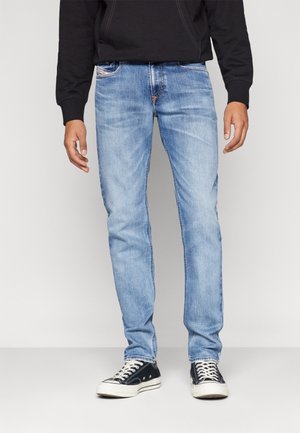 Mann trägt hellblaue Slim-Fit-Jeans und schwarze Low-Top-Sneaker, steht vor einem schlichten, hellgrauen Hintergrund.