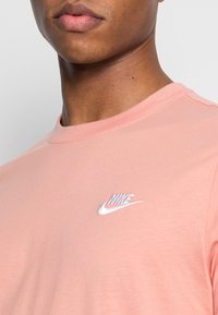 Světle růžové bavlněné tričko s kulatým výstřihem a bílým vyšitým logem Nike na levé straně hrudi. Hladký povrch.