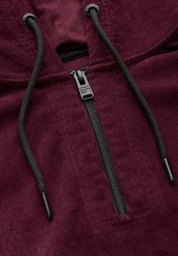 Burgundy huvtröja med halv dragkedja fram, svarta snörningar och en texturerad tyg. Har en logobroderad dragkedja.