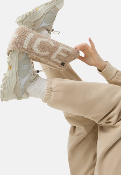 Beige strikket lægvarmer med "ICE" mønster, båret over en chunky hvid sneakers med grå mesh og detaljer, parret med beige joggerbukser.