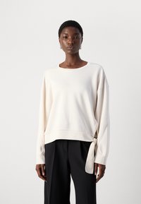Lichtbeige oversized sweatshirt met een ronde hals, verlaagde schouders en een strikdetail aan de onderkant. Zachte, gladde textuur van de stof.