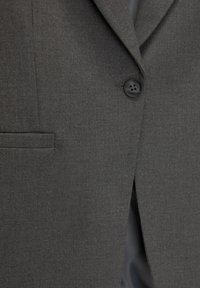 Blazer gris sur mesure en tissu texturé, doté d'un unique bouton noir et d'une poche poitrine. Lignes épurées et design minimaliste.