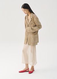 Blazer en suédine beige avec deux poches avant, superposé à une robe beige semi-transparente. Escarpins rouges à enfiler avec une semelle plate et un bout pointu.