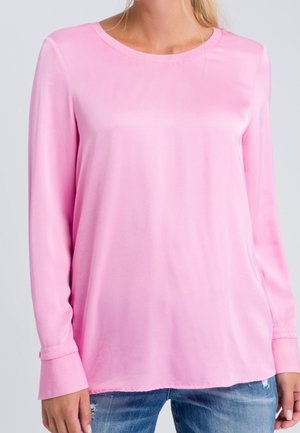 Femme portant un chemisier à manches longues, rose clair, ample, associé à un jean bleu délavé sur un fond uni.