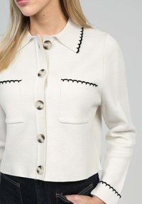 Cardigan en tricot blanc avec un col pointu, des coutures noires en feston le long des coutures, et de grands boutons. Deux poches poitrine sont visibles.