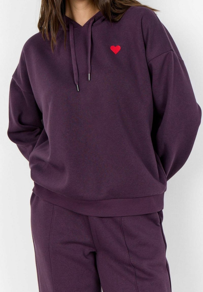 Femme portant un sweat à capuche violet foncé et un pantalon assorti avec un petit emblème de cœur rouge sur la poitrine gauche, debout sur un fond clair.