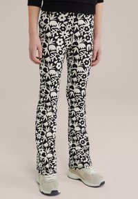 Fekete, harangszárú leggings, fehér virág- és levélmintákkal. Rugalmas anyag, közepes derék, részletek a derékszíjon. Világos tornacipővel párosítva.