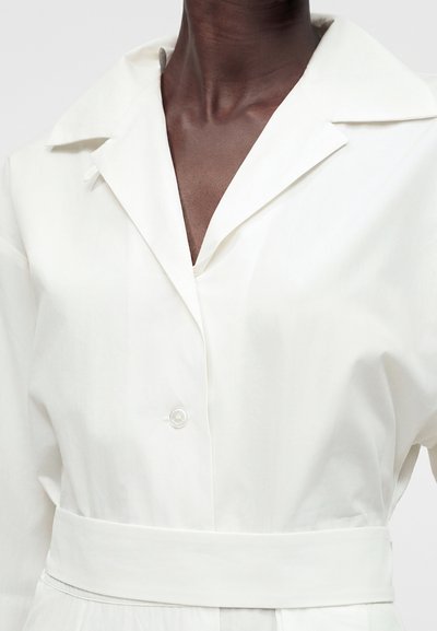 Άτομο με σκοτεινό δέρμα φοράει λευκό πουκάμισο με γιακά, φαρδιά ζώνη και ένα κουμπί κλεισμένο στο στήθος.