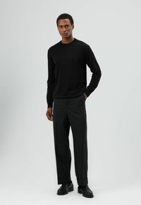 Zwarte trui met lange mouwen gecombineerd met antracietgrijze broek met krijtstrepen en zwarte leren schoenen. Simpel, aansluitend ontwerp met minimale details.