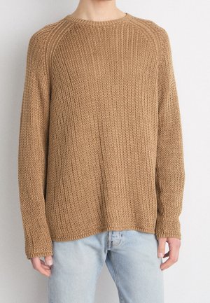 Person trägt einen locker sitzenden, langärmligen, hellbraunen Strickpullover und hellblaue Jeans und steht vor einem schlichten Hintergrund.