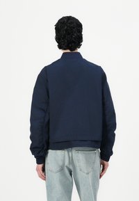 Mørkeblå bomberjakke med ribbestrikket krage og mansjetter, med minimalistisk design og glatt tekstur. Brukt med lyseblå jeans.