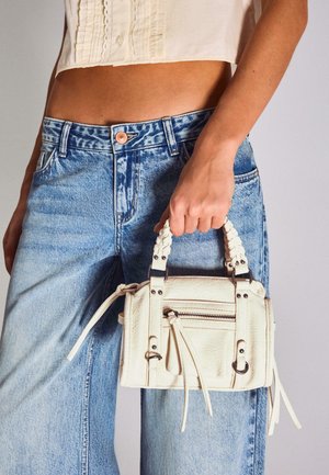 Žena v světle modrých džínách a krémovém krátkém topu držící malou bílou koženou kabelku s pletenými úchyty a zipovými detaily.