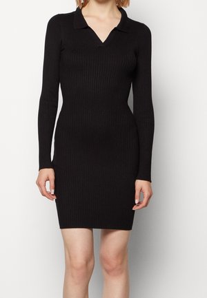 Kobieta ubrana w dopasowaną, czarną, prążkowaną sukienkę z długim rękawem, kołnierzykiem i głębokim dekoltem w kształcie litery V, stojąca na białym tle.