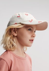 Hvit baseballcaps med røde kirsebær- og ankertrykk, buet skygge, i bomull. Lyst t-skjorte med rund hals.