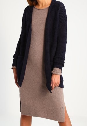 Cardigan de punto azul marino con apertura frontal, llevado sobre un vestido ajustado de color marrón claro. Presenta puños acanalados y aberturas laterales para mayor comodidad al moverse.