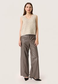 Top senza maniche beige texturizzato con scollo a V, abbinato a pantaloni larghi in pelle marrone a vita alta e décolleté neri con punta appuntita.