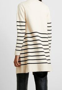 Jasno beżowy kardigan z czarnymi poziomymi paskami, długimi rękawami, prostym krojem i rozcięciem na dole. Miękka faktura, minimalistyczny design.