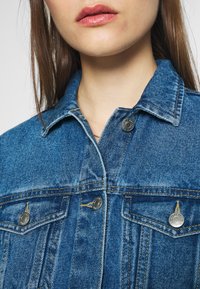 Denim jacket in medium blue, featuring a classic collar, silver snap buttons, and two chest pockets with visible stitching details.