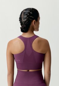 Lila träningssport-bh med racerback-design, strukturerad tyg och en åtsittande form. Har en kontrasterande band och subtil logotyp.