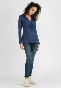 Marineblaues Langarm-Wickeltop mit geraffter Taille, kombiniert mit blauen Slim-Jeans und hellen Sneakers. Weicher, elastischer Stoff.