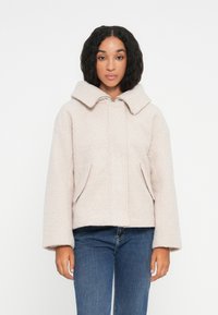 Lys rosa cropped jakke laget av mykt, teksturert stoff med bred krage, store lommer og en avslappet passform. Kombinert med blå jeans.