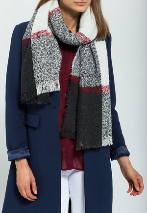 Femme portant une écharpe en tricot épaisse multicolore sur un manteau bleu marine, une chemise bordeaux boutonnée et un pantalon blanc, vue du cou aux hanches.