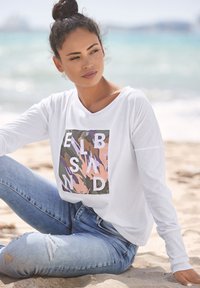 Weißes Langarmhemd mit einem bunten Grafikdesign und Buchstaben auf grünem Hintergrund; kombiniert mit hellblauen, ausgebeulten Jeans.