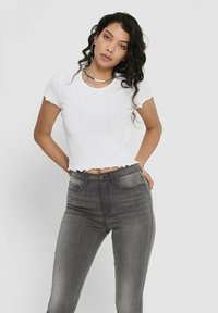White cropped ribbed top with scalloped edges, paired with high-waisted gray jeans. Model has curly hair and wears hoop earrings.