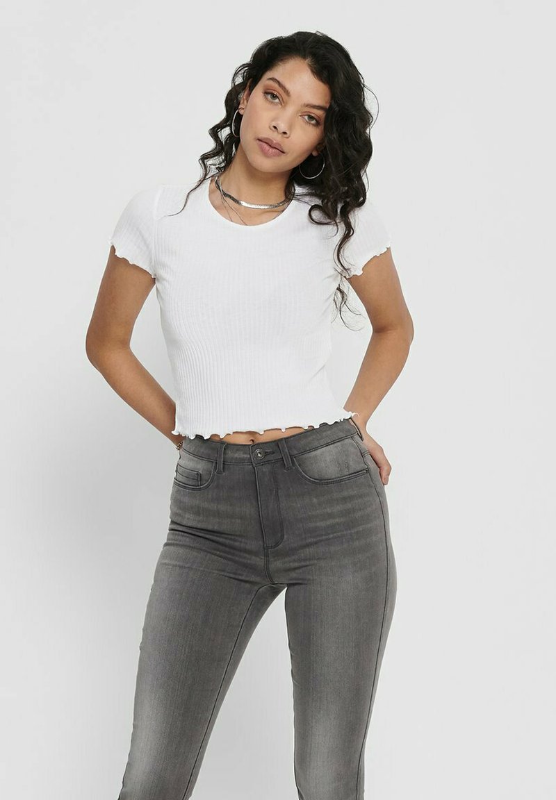 White cropped ribbed top with scalloped edges, paired with high-waisted gray jeans. Model has curly hair and wears hoop earrings.