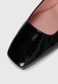 Černá lakovaná bota s čtveratou špičkou a růžovým vnitřním lemováním, zobrazená zblízka z vrchního úhlu na bílém pozadí.