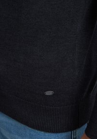 Ciemnoniebieski sweter dziany z prążkowanym wykończeniem, z małym metalowym logo na dolnej stronie. Materiał wydaje się być miękki i teksturowany.