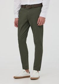 Pantaloni khaki a gamba affusolata realizzati in cotone, con una texture liscia, una cintura marrone e abbinati a sneaker bianche, su uno sfondo neutro.