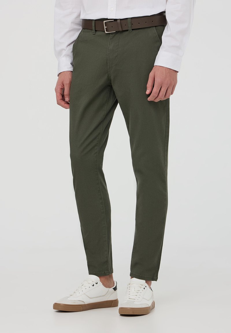 Pantaloni khaki a gamba affusolata realizzati in cotone, con una texture liscia, una cintura marrone e abbinati a sneaker bianche, su uno sfondo neutro.