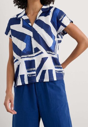 Vrouw draagt een shirt met korte mouwen met een blauw-wit abstract patroon en effen blauwe broek, staand met één hand in de zij.