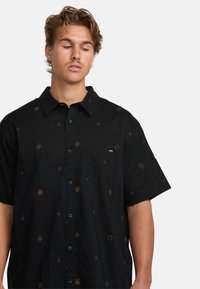 Camisa negra de botones con mangas cortas, con patrones de bordado geométrico dorado y un bolsillo en el pecho. Hecha de tejido ligero.