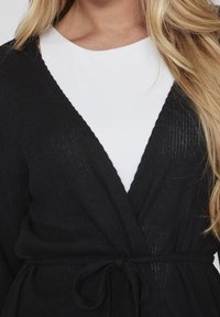 Černý žebrovaný pletený cardigan s ovínacím designem, zavazováním v pase a hlubokým výstřihem do V. Přeložený přes bílý top, ukazuje kontrastní barvy.