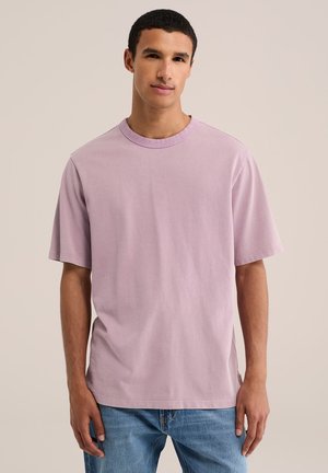 Młody mężczyzna z krótkimi kręconymi włosami, ubrany w jednolity jasnofioletowy t-shirt z krótkim rękawem i niebieskie dżinsy, stojący na neutralnym tle.