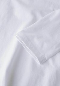 Biała bawełniana koszulka o miękkiej teksturze, z zamkniętym rękawem i gładkim, przeszytym szwem. Prosty design bez widocznych wzorów czy elementów metalowych.