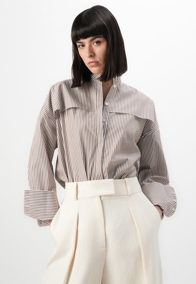 Blusa a righe beige e marrone con colletto alto e chiusura frontale a bottoni, abbinata a pantaloni plissettati crema con vita larga.