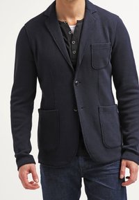 Blazer blu navy realizzato in tessuto testurizzato con due tasche frontali, revers a scollar, e chiusura a un bottone, indossato sopra una camicia nera.