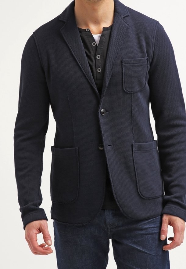 Blazer blu navy realizzato in tessuto testurizzato con due tasche frontali, revers a scollar, e chiusura a un bottone, indossato sopra una camicia nera.