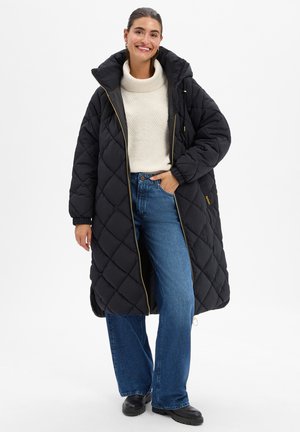 Vrouw met een lange zwarte doorgestikte jas over een witte coltrui, blauwe spijkerbroek en zwarte laarzen, glimlachend met één hand in haar zak.