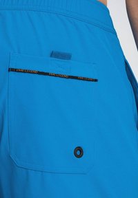 Shorts de bain bleus avec une poche arrière, dotés d'un bouton noir et d'une étiquette décorative noir et jaune portant la mention "PME LEGEND." Texture lisse.