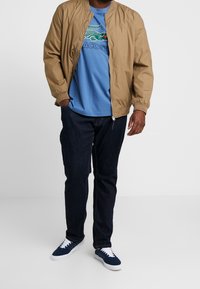 Kurtka typu bomber w kolorze piaskowym, niebieska koszulka z nadrukiem krokodyla, ciemnoniebieskie dżinsy i granatowe trampki z białymi sznurowadłami. Codzienny strój.