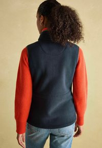 Námořnická modrá fleecová vesta s vysokým límcem, skombinovaná s oranžovým žebrovaným svetr. Má přiléhavý střih a viditelné švy.