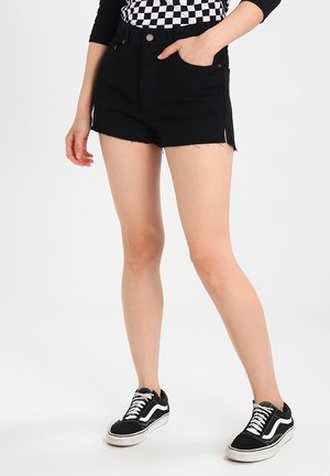 Pantalones cortos de mezclilla negros con una cintura alta, dobladillo deshilachado y espacios laterales; combinados con zapatillas en blanco y negro. Parte superior de patrón a cuadros visible.