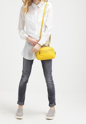 Keltainen nahkainen olkalaukku, jossa on sileä pinta, kaksi vetoketjullista osastoa ja säädettävä hihna. Yhdistetty valkoiseen paitaan ja harmaisiin farkkuihin.