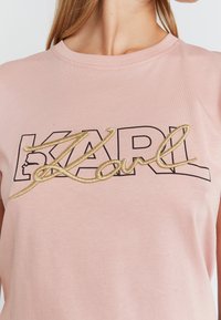 Vaaleanpunainen puuvillainen t-paita, jossa on lyhyet hihat ja musta "KARL"-printti sekä kultakirjailtu allekirjoitusyksityiskohta. Sileä pinta.