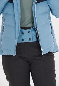 Lyseblå isoleret jakke med høj krave, frontlynlås og sorte detaljer. Sorte bukser med en tætsiddende pasform og en knaplukning i taljen.