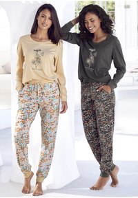 Dos mujeres llevan conjuntos de ropa de estar por casa. A la izquierda: camiseta crema con estampado de "FLORES SILVESTRES", pantalones floreados. A la derecha: camiseta gris oscuro, pantalones oscuros con flores.